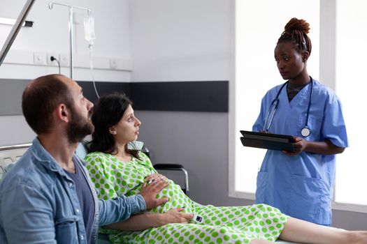 African american nurse monitoring healthcare of pregnant woman sitting in hospital ward bed. Adult with pregnancy and father of child waiting on baby delivery procedure at maternity