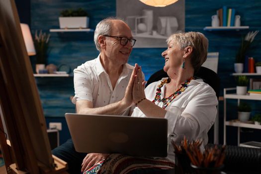 Aged couple of artists highfive while using laptop for design