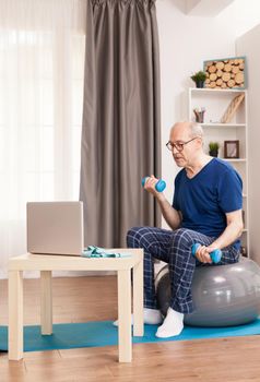 Senior man training biceps watching online fitness lesson. Old person pensioner online internet exercise training at home sport activity with dumbbell, resistance band, swiss ball at elderly retirement age.