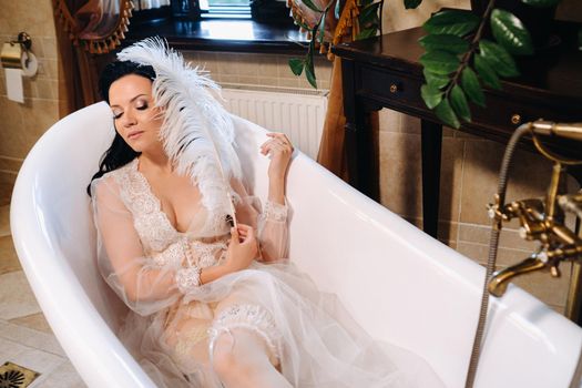 the bride, dressed in a boudoir transparent dress and underwear, lies in a vintage bathroom with a white feather in her hands, The morning of the bride