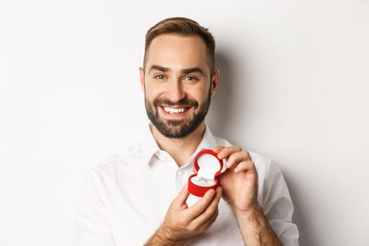 Close-up of happy handsome man making a proposal, holding wedding ring in box and smiling, asking to marry him, white background