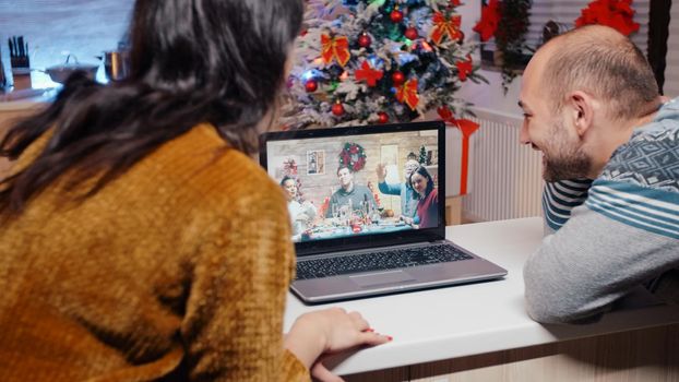 Man and woman using video call communication talking to family on christmas eve. Couple chatting with relatives on online remote conference, celebrating holiday festivity in decorated kitchen.
