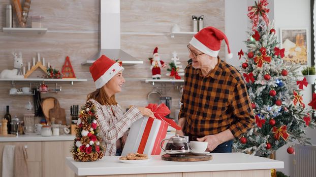 Grandfather surprising granddaughter with wrapper present gift in xmas decorated kitchen