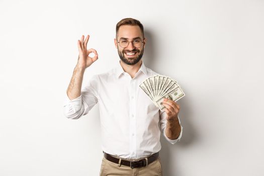 Satisfied young businessman showing money, make okay sign, earning cash, standing over white background