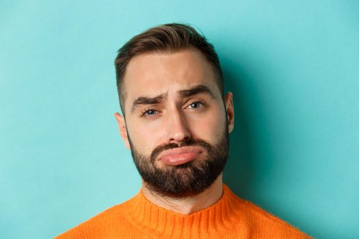 Headshot of sad and gloomy man complaining, pouting and frowning disappointed, standing against turquoise background