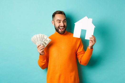 Real estate concept and mortgage concept. Cheerful man holding money and paper house, smiling excited, buying or renting flat, standing over blue background