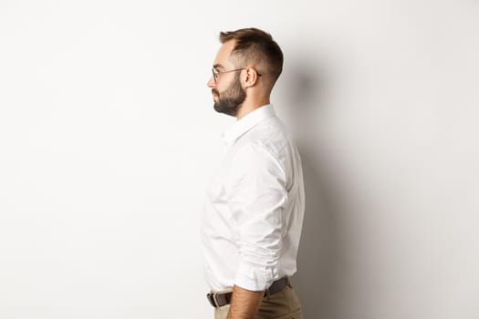 Profile of young businessman in white collar shirt and beige pants, looking left, standing against studio background