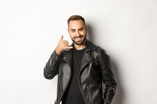 Handsome man in leather jacket flirting with you, showing mobile call me gesture, standing against white background