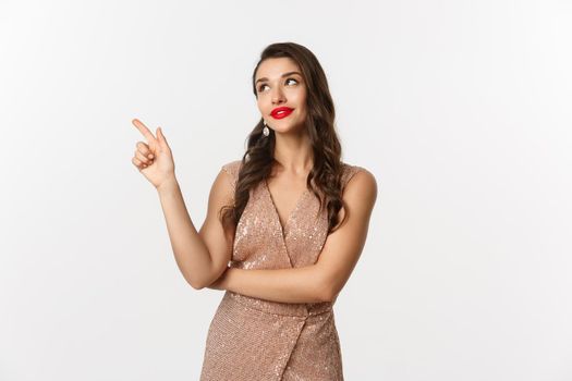 Christmas, holidays and celebration concept. Dreamy beautiful woman in party dress, pointing and looking left, thinking about new year party, white background