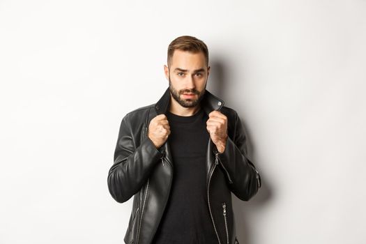 Image of handsome and confident man putting on leather biker jacket, standing against white background