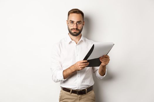 Serious employer looking at camera while reading documents on clipboard, having job interview, standing over white background