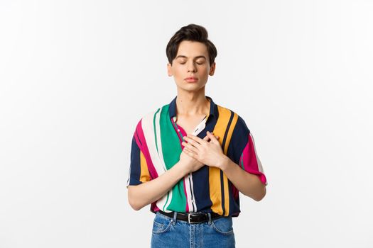 Image of nostalgic young gay man standing with closed eyes and hands on heart, remember something, standing over white background