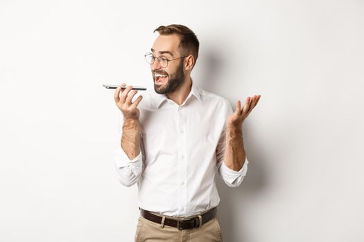 Excited businessman talking on speakerphone and smiling, record voice message with ecstatic face, standing over white background