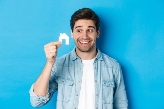 Real estate concept. Excited guy looking at small house model and smiling, renting apartment, standing over blue background