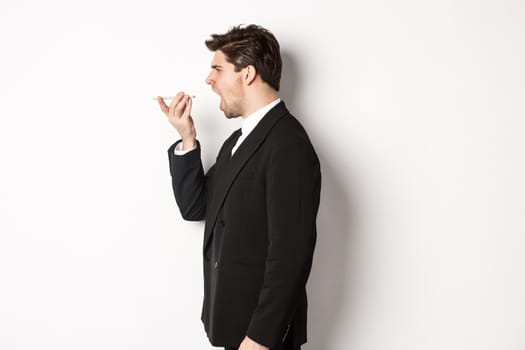 Profile shot of angry businessman in black suit, shouting at speakerphone and looking mad, recording voice message, standing over white background