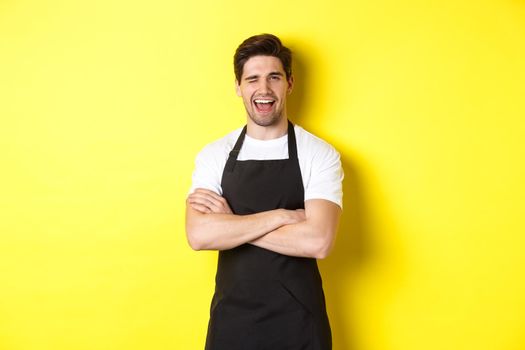 Handsome barista in black apron winking at you, wearing black apron uniform, standing over yellow background