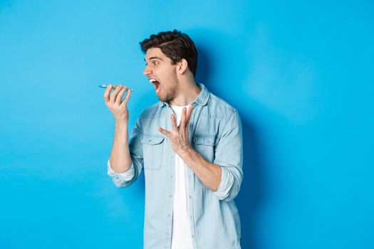 Angry and annoyed guy screaming at mobile phone, shouting on speakerphone while standing over blue background