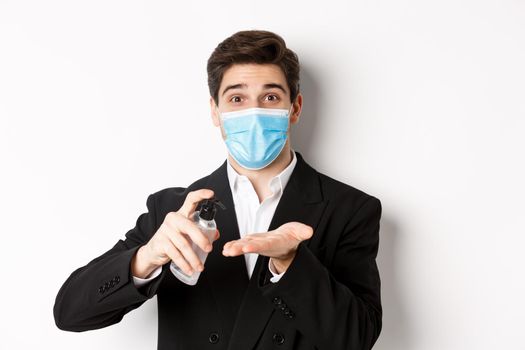 Concept of covid-19, business and social distancing. Image of handsome businessman in trendy suit and medical mask, cleaning hands with hand sanitizer, standing over white background