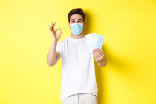 Concept of covid-19, quarantine and preventive measures. Satisfied man showing okay sign and giving medical masks, standing over yellow background