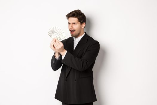 Image of confused businessman looking at fake money, standing over white background in black suit