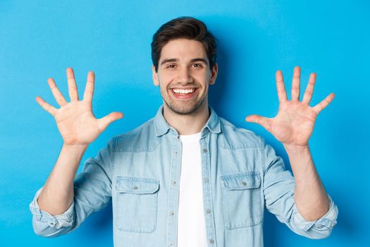 Close-up of handsome man smiling, showing fingers number ten, standing over blue background