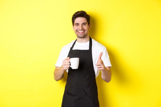 Barista bringing coffee and pointing finger gun at camera, standing in black apron against yellow background