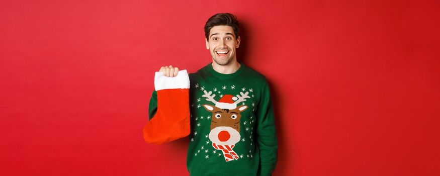Image of handsome happy guy in sweater, holding christmas stocking and smiling amused, standing against red background