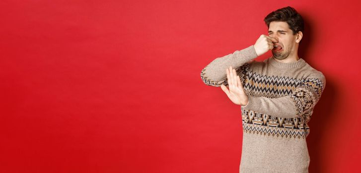 Image of disgusted handsome man in winter sweater, rejecting something with bad smell, shut nose and raising hand to decline, standing over red background.