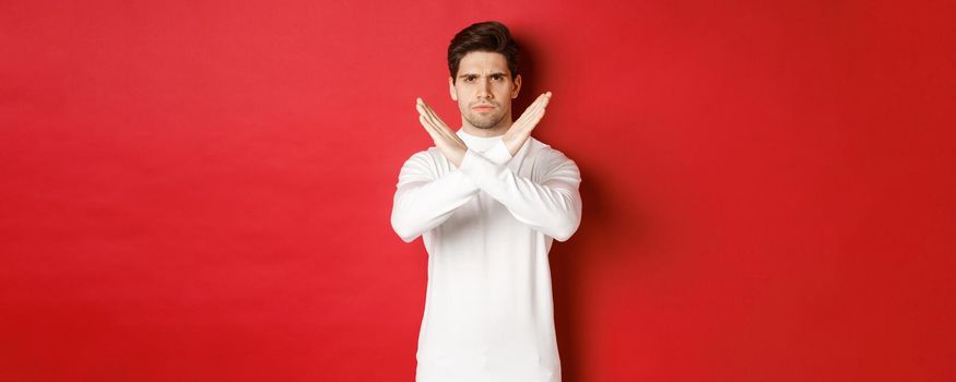 Image of serious and angry man in white sweater, express storng disapproval, showing cross sign to stop something bad, forbid action, standing over red background
