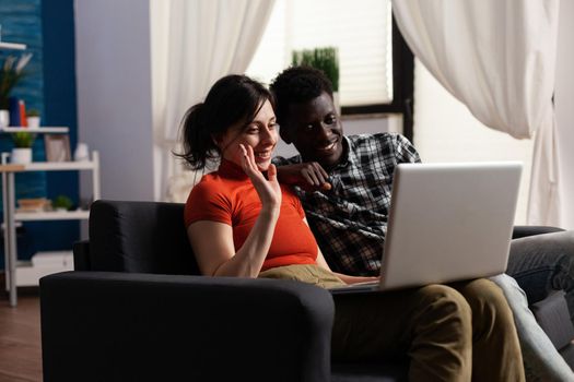Interracial couple using video call communication on laptop at home. Mixed race people waving at camera while talking to friends on online remote conference via internet connection