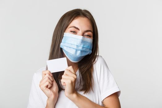 Covid-19, health and social distancing concept. Close-up of attractive happy brunette girl in medical mask showing credit card.
