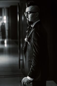 The groom in a tuxedo and bow tie with a mohawk hairstyle and a cane in the interior. black and white photo