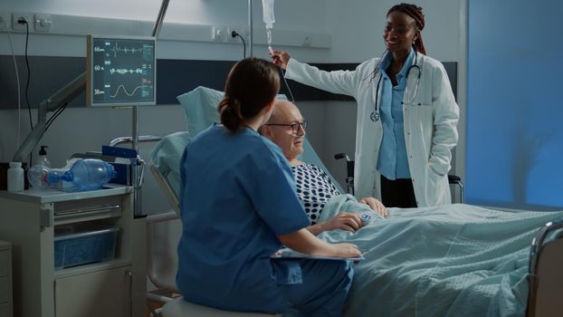 Multi ethnic medical staff checking patient healthcare problems to use healing treatment. Nurse and african american doctor tend to sick old man on bed in modern hospital ward