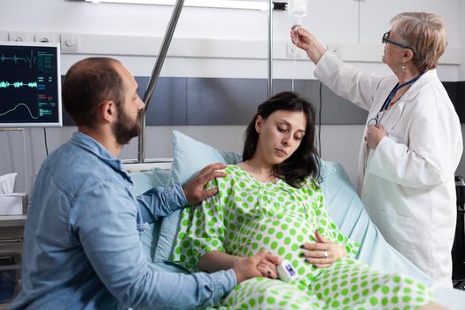Doctor checking healthcare of pregnant woman in hospital ward with father of child supporting and holding hand. Couple expecting baby receiving examination from obstetrician at maternity