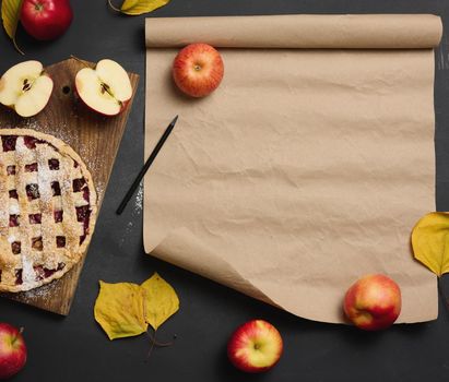 baked round traditional apple pie on a brown wooden board and fresh red apples. Nearby is a roll of brown paper for writing a recipe or menu, top view