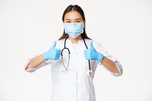 Healthcare and covid-19 preventive measures concept. Asian physician in face mask and rubber gloves, shows thumbs up, stands in uniform over white background