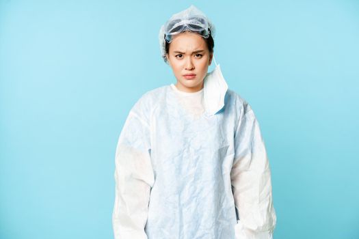 Angry asian woman doctor or nurse, wearing personal protective equipment, looking with disapproval, furrow eyebrows upset, standing over blue background