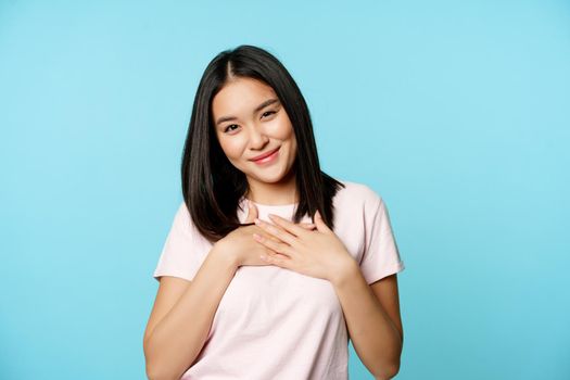Smiling asian female model holds hands on heart, cherish smth, feeling care and warm love feelings, standing in t-shirt over blue background