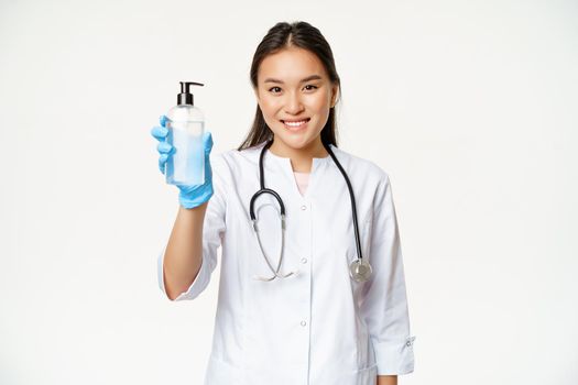 Smiling asian doctor showing hand sanitizer, bottle of antiseptic, wearing sterile rubber gloves and clinic uniform, white background