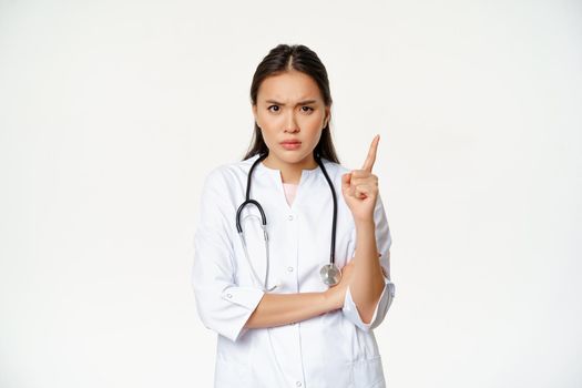 Doctor shows disapproval, scolding patient. Angry female asian physician shakes finger and furrow eyebrows, disappointed with bad behaviour, standing over white background