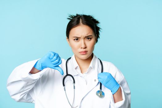 Angry asian woman doctor, physician in uniform and gloves shows thumbs down, furrow eyebrows upset, disapprove smth, blue background