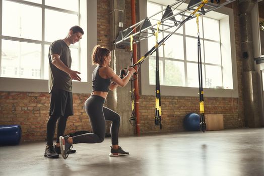 Postive workout. Beautiful young woman in sportswear doing trx exercises at gym while her personal trainer is standing next to her. TRX Training. Exercising together. Active lifestyle