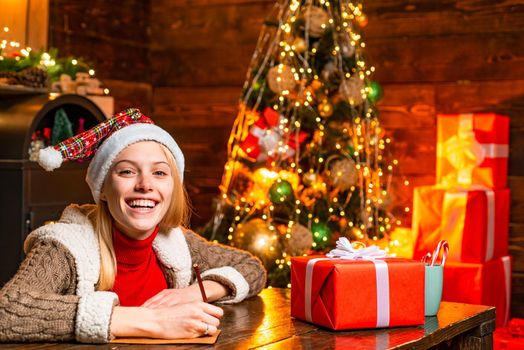 Cheerful smiling blonde woman writing a letter to Santa with her wish list of presents. Christmas presents gifts concept. Joy and happiness. Christmas. Xmas tree.