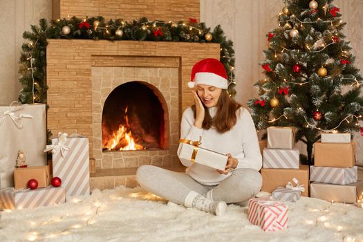 Beautiful young woman with dark hair, smiling, holding festive gift in hands while sitting near fireplace and Christmas tree. Happy girl dressed in santa hat, warm sweater and socks.