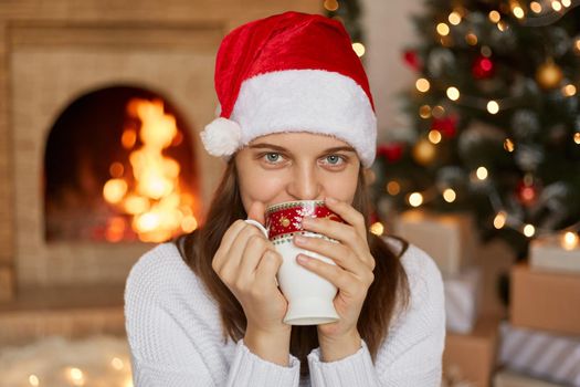 Beautiful young woman wearing Santa Claus red hat sitting near fire place in living room with x-mas decoration, lady drinking hot beverage, holding cup with both hands, looks at camera.
