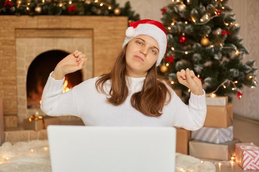 Beautiful woman sitting on floor working with laptop at home around christmas tree, stretching her back, tired and relaxed, girl wearing white sweater and santa claus hat.