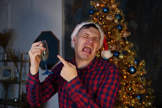 Young man in santa hat drinks champagne and grimaces from alcohol on background of Christmas tree in dark room. Concept of Christmas celebration at home.