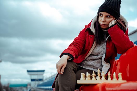 Young woman in winter clothes plays chess, sitting on stadium bleachers alone. Female in black cap with chess on sports stadium in cloudy weather.