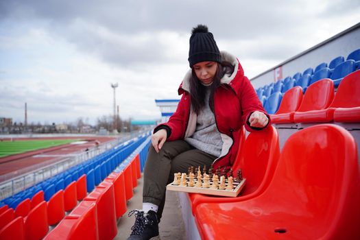 Young woman in winter clothes plays chess, sitting on stadium bleachers alone. Female in black cap with chess on sports stadium in cloudy weather.