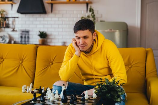 Young man sitting on yellow sofa and playing chess in room. Male playing in logical board game with himself.